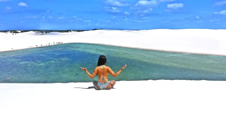 Turista sentada na areia branca diante de uma lagoa verde-azulada